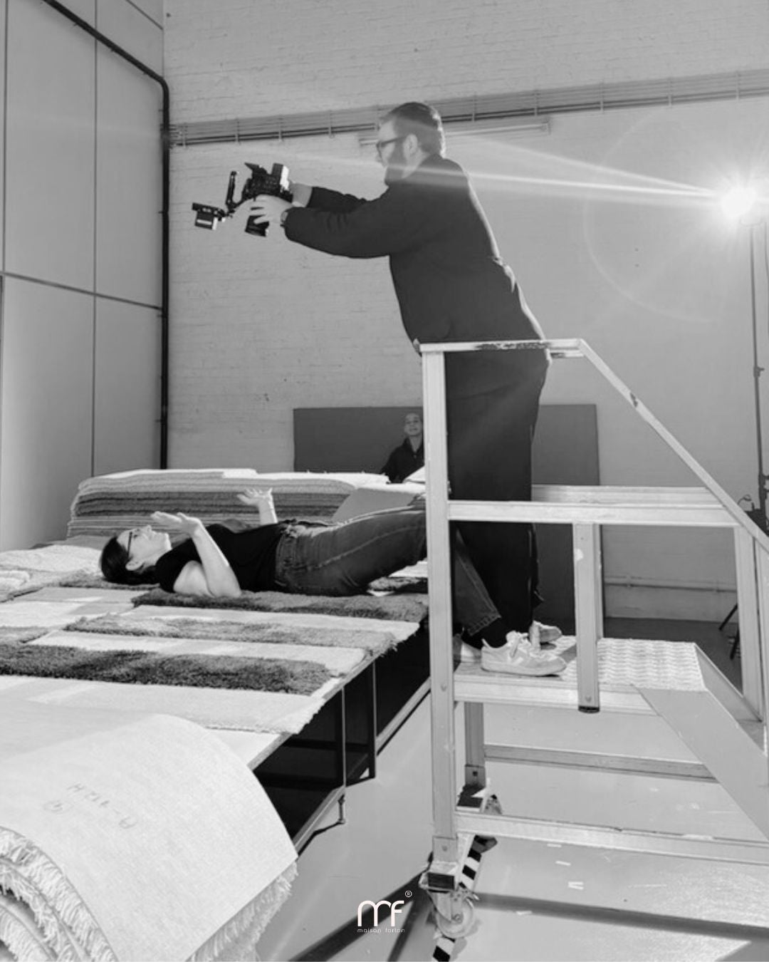 Cameraman maakt een opname van bovenaf terwijl Jozefien tussen tapijtstalen op een tafel ligt; hij staat op een kleine trap voor een beter perspectief in een atelieromgeving.