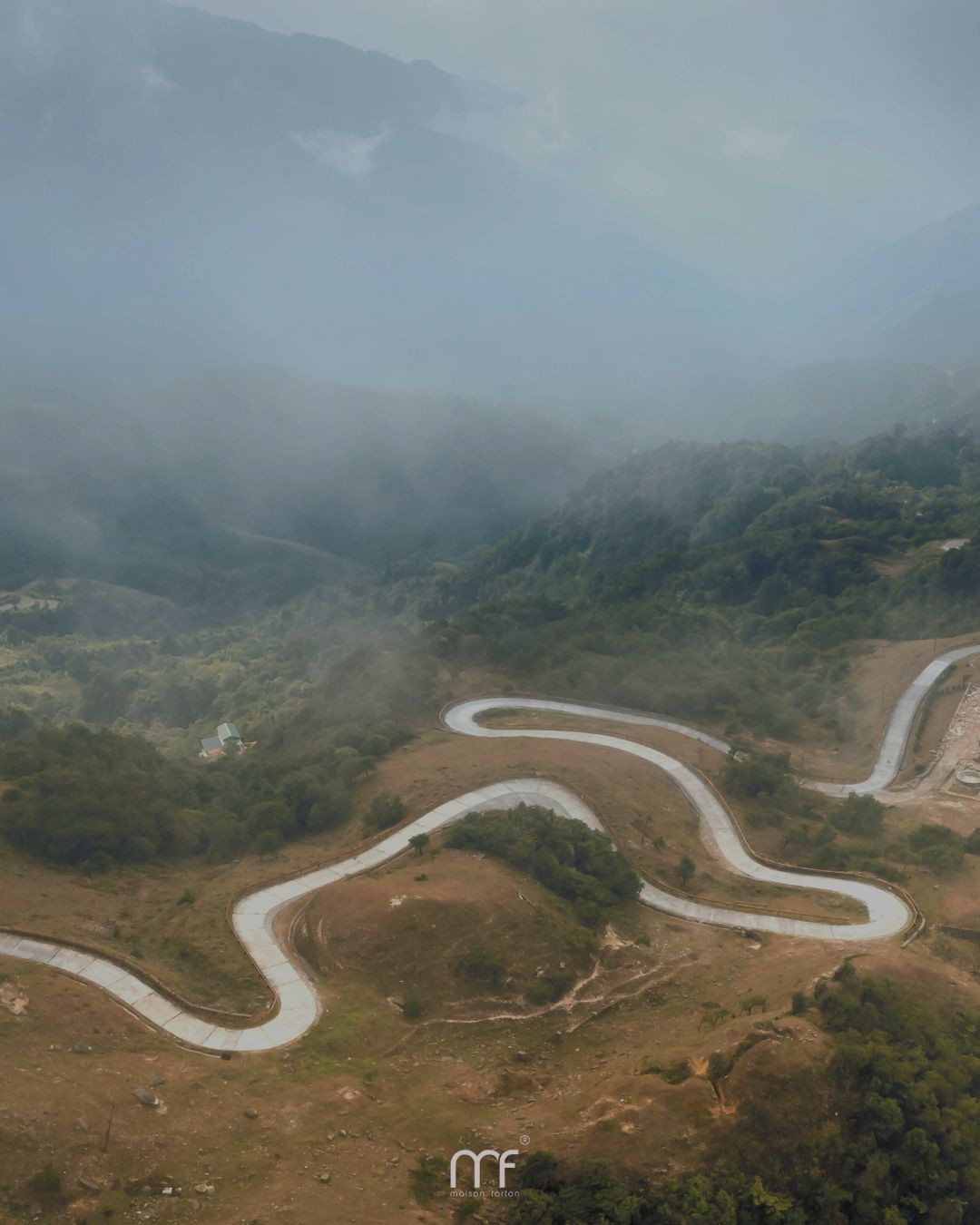 Luchtfoto van een kronkelende bergweg die door groen heuvellandschap loopt, omringd door mist die langzaam over de valleien en bergflanken hangt.