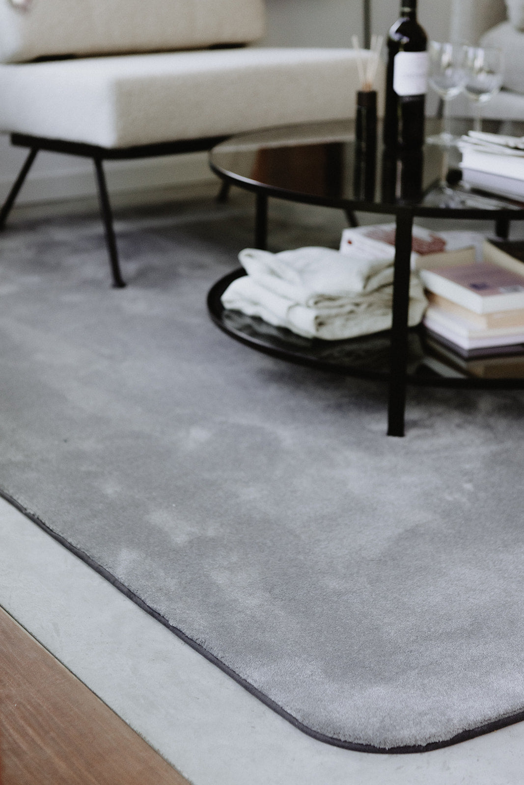 Detail ronde hoek van duurzaam grijskleurig Rounded01. tapijt met stone kleurrand, met op de achtergrond een zetel en salontafel.