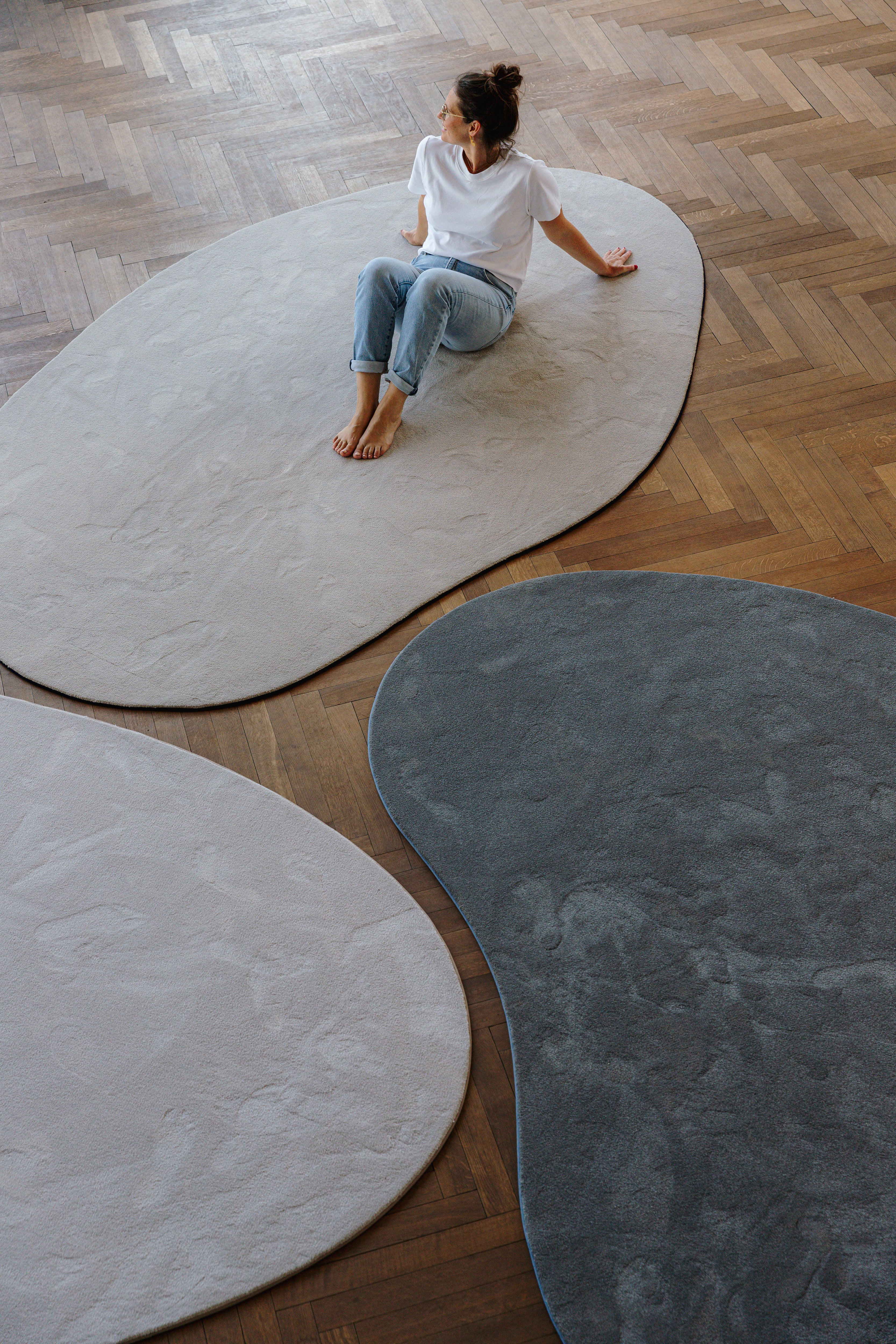 Vrouw (Jozefien Forton) zittend op collectie organische vormen tapijten Belgian soft design op parket vloer.